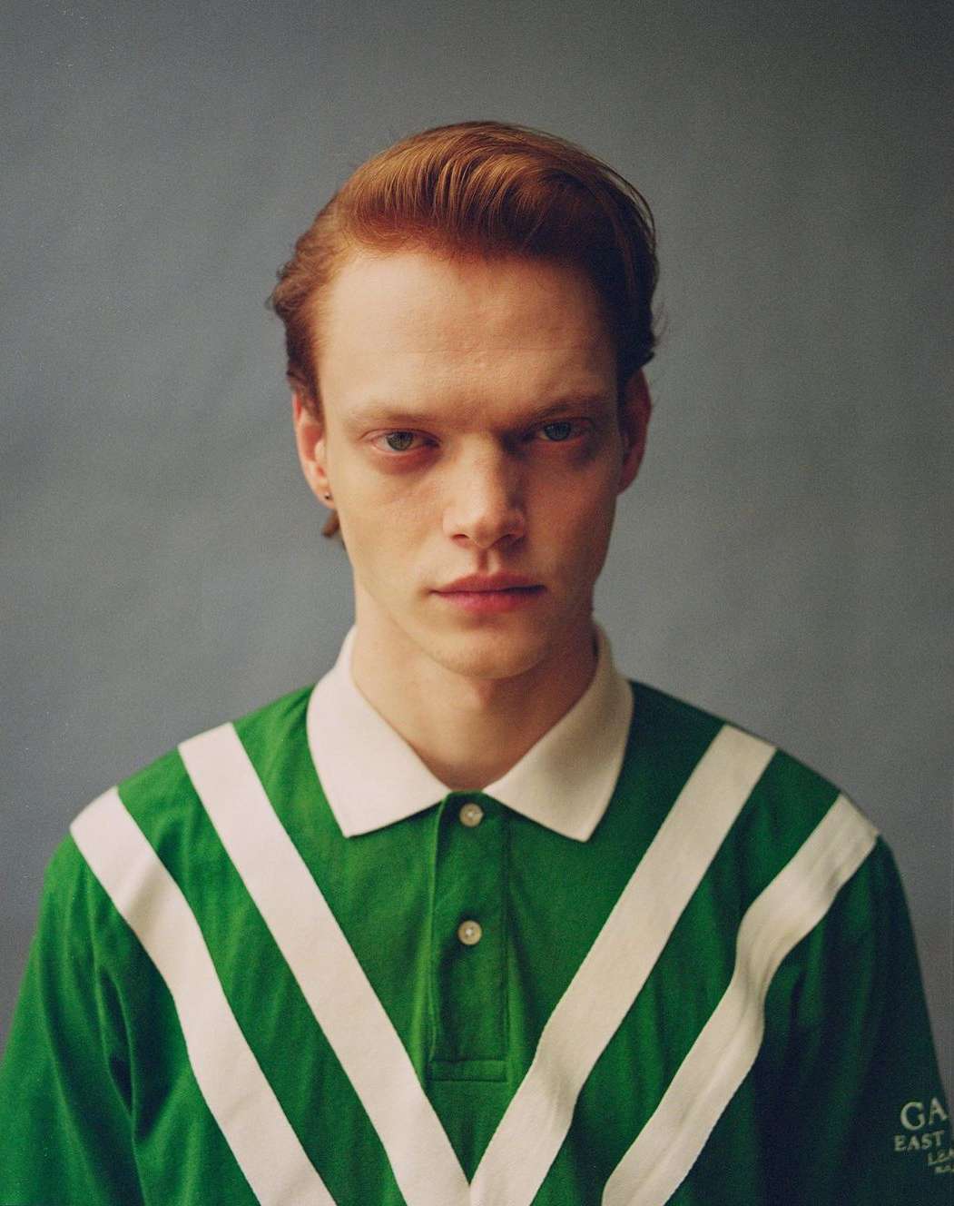 Portrait of a red-haired man wearing a green polo shirt with white stripes, against a gray background in a studio setting.