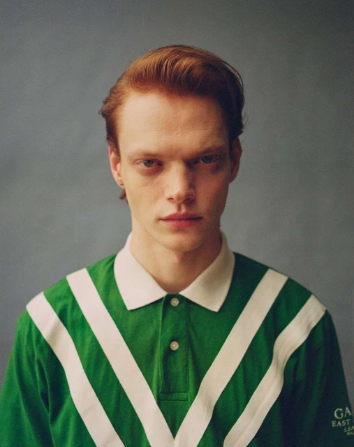 Portrait of a red-haired man wearing a green polo shirt with white stripes, against a gray background in a studio setting.