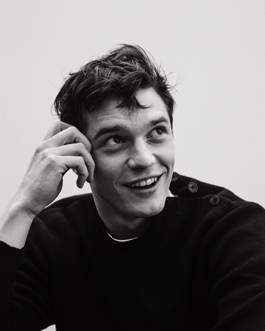 Monochrome portrait of smiling adult male with dark hair, head resting on hand, wearing a crewneck sweater and looking upward.