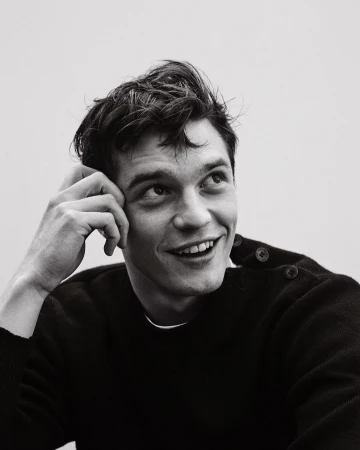 Monochrome portrait of smiling adult male with dark hair, head resting on hand, wearing a crewneck sweater and looking upward.