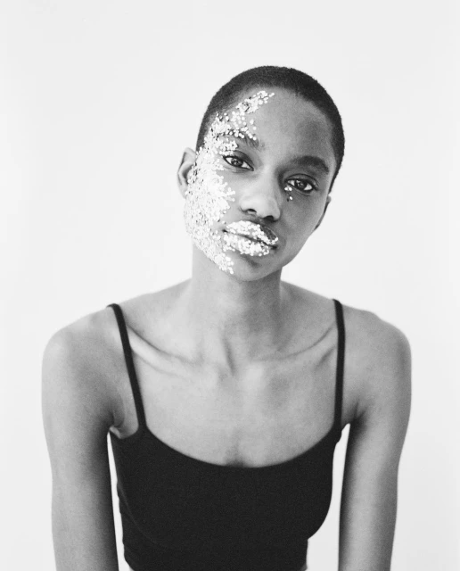 Striking black and white portrait of a Black woman with glitter on face and lips against plain white background, creating stark contrast.