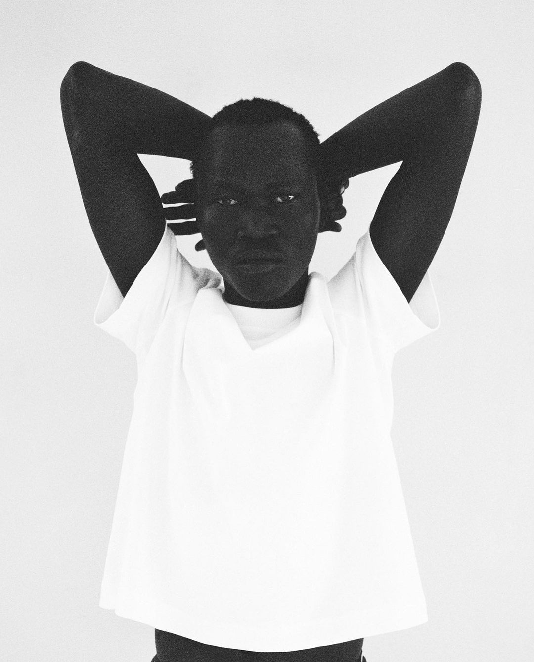 Close-up black and white portrait of an adult woman with her hands behind her head, wearing a white shirt in a neutral setting.