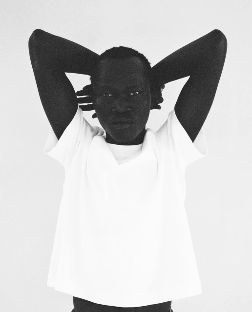 Close-up black and white portrait of an adult woman with her hands behind her head, wearing a white shirt in a neutral setting.