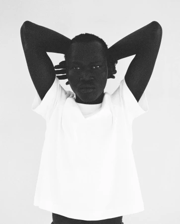 Close-up black and white portrait of an adult woman with her hands behind her head, wearing a white shirt in a neutral setting.