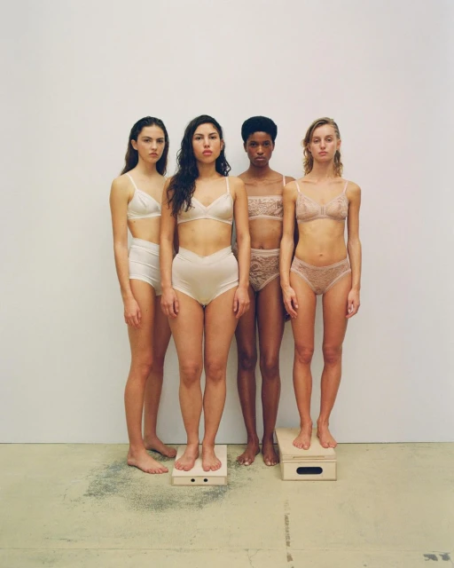 Four women stand in lingerie, posed against a stark white backdrop with bare feet on wooden blocks, showcasing different skin tones and body types.