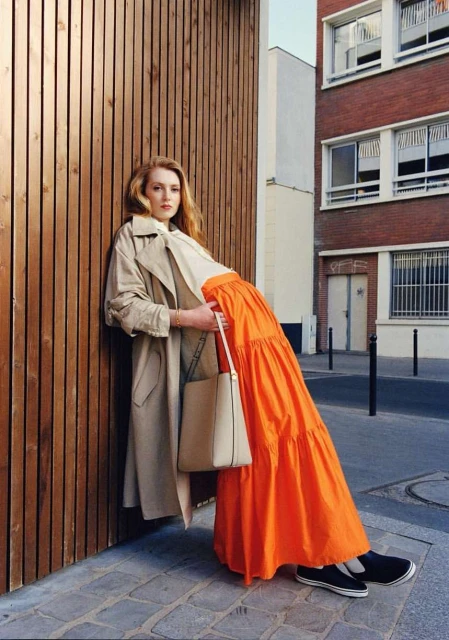 Stylish woman in trench coat and vibrant orange skirt poses against wood and brick walls in an urban setting, showcasing fashion.