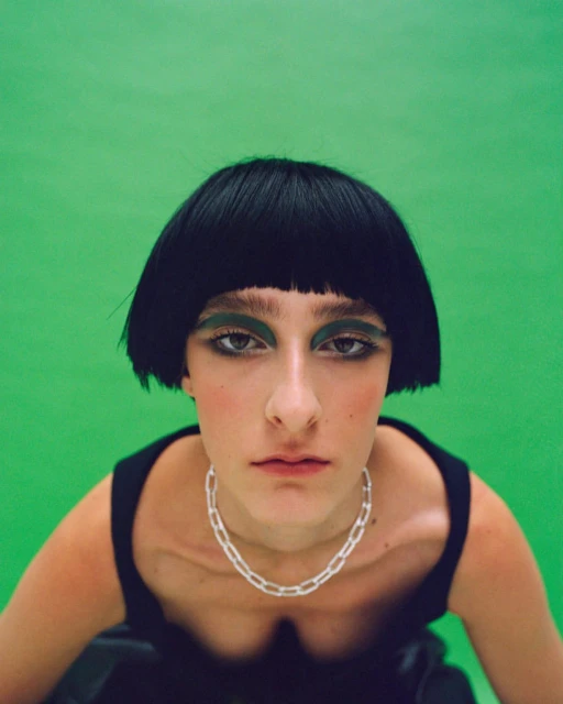 Woman with a bob hairstyle and heavy eye makeup in a black sleeveless top in a studio with a green background looking at the camera.