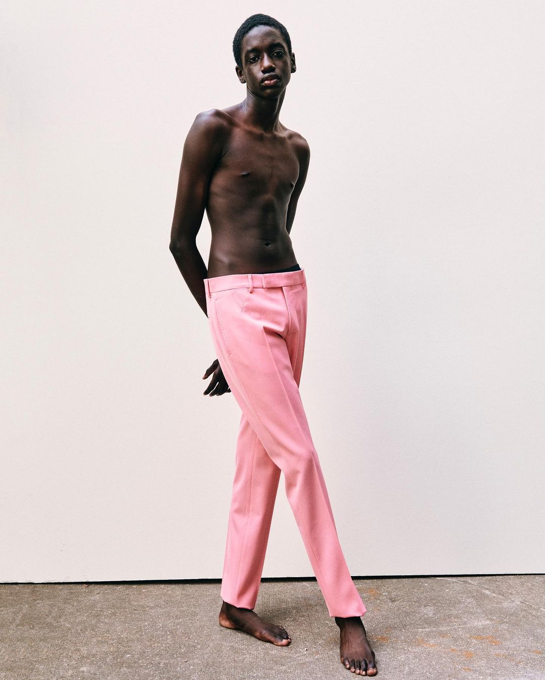 Full portrait of a slender adult male with dark skin, posing shirtless in light-pink trousers against a white studio background.