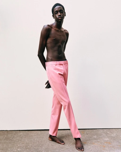 Full portrait of a slender adult male with dark skin, posing shirtless in light-pink trousers against a white studio background.