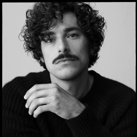Portrait of a man with curly hair and a mustache wearing a dark sweater against a seamless light background by Arthur Delloye.