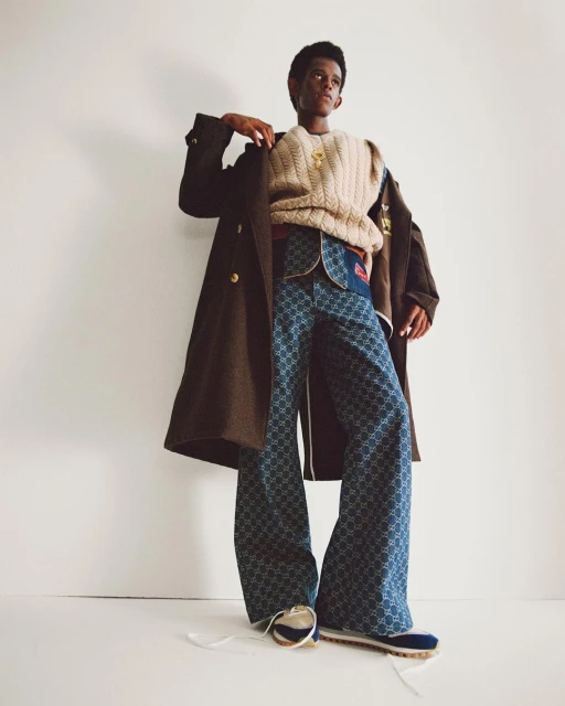 A person in patterned blue pants, tan sweater vest, and a brown coat stands in front of a white background in a fashion pose.