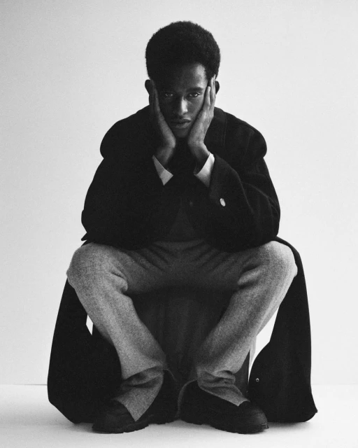 Black and white portrait of a young adult male in a coat and pants seated with his hands on his face against a light backdrop.