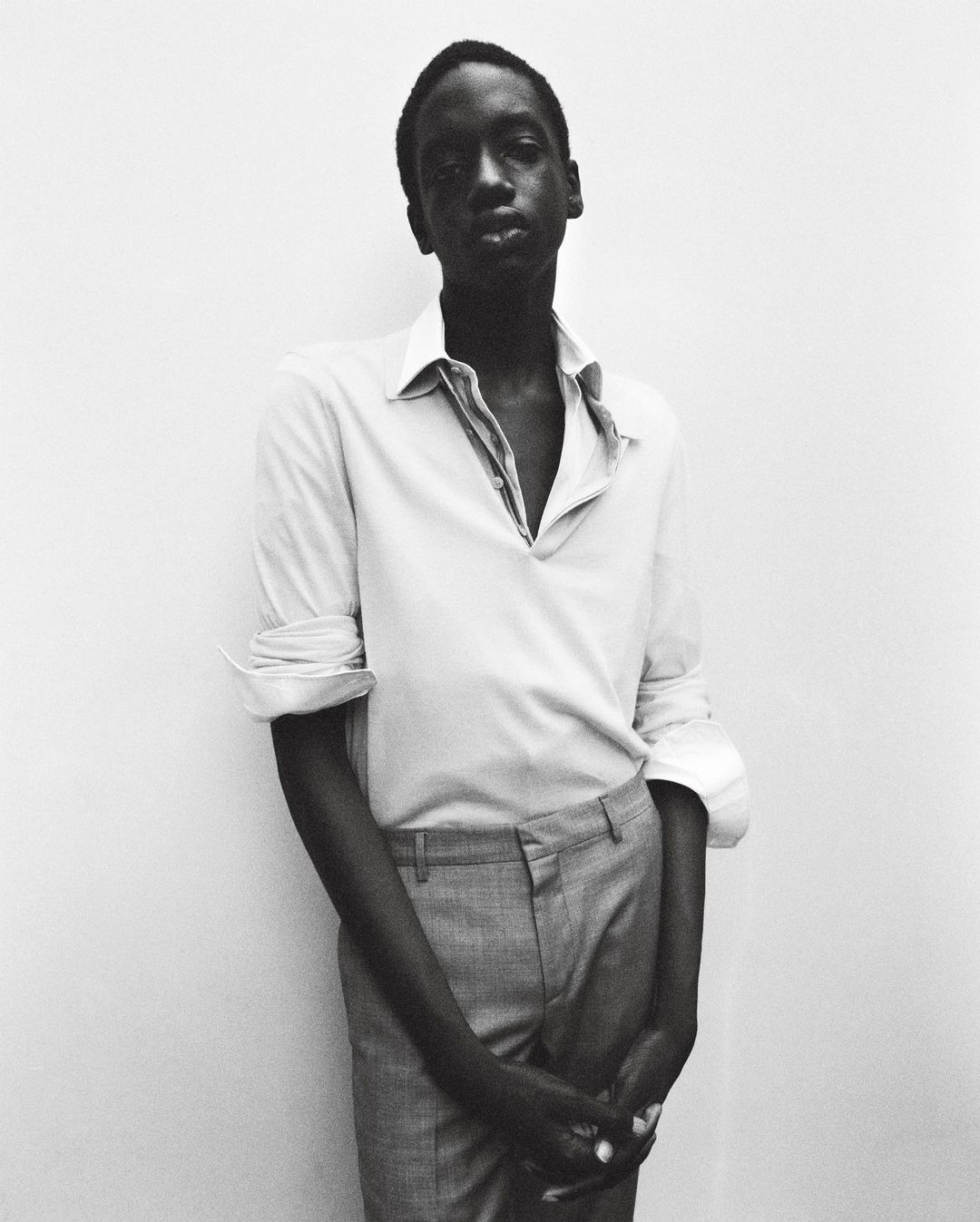 Monochromatic portrait of an African man with an open-collar shirt and grey pants, standing against a light wall. Fashion and style.