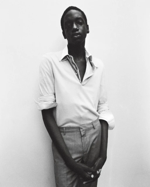 Monochromatic portrait of an African man with an open-collar shirt and grey pants, standing against a light wall. Fashion and style.