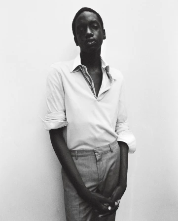 Monochromatic portrait of an African man with an open-collar shirt and grey pants, standing against a light wall. Fashion and style.
