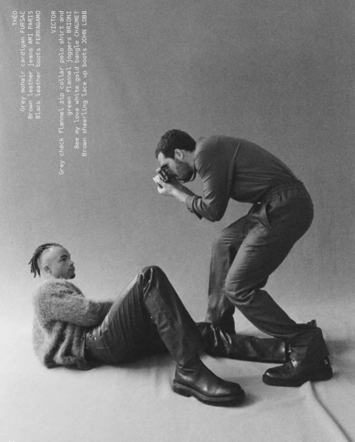 Two adult males in a photo studio: one lying down wearing leather pants and boots, the other standing taking a photo, gray background.