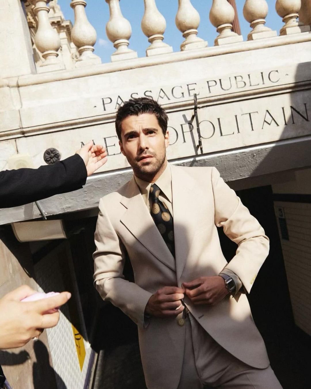 A stylish man in a beige suit poses near a 'Passage Public' sign, exuding confidence and urban charm with classic fashion elements.