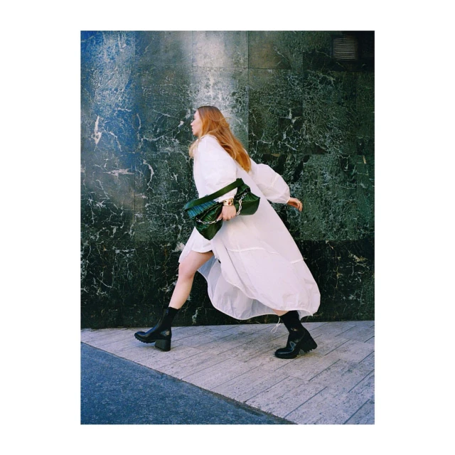 A tall adult woman walks on a sidewalk in an elegant white dress, dark boots, and a green handbag in an outdoor fashion shoot.