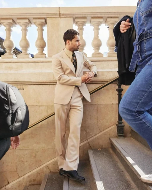 Stylish man in a beige suit stands on outdoor stairs, looking away. People walk past him, and a stone balustrade is in the background.