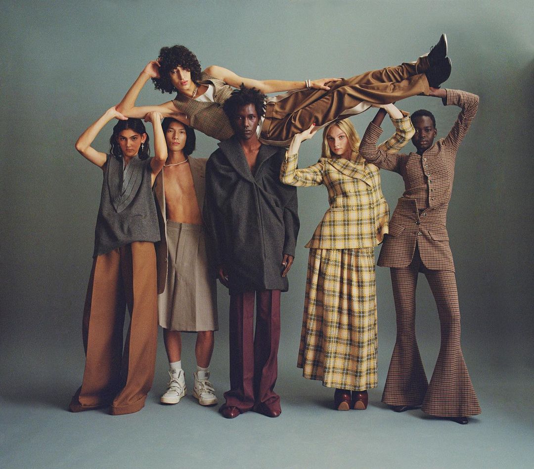 Seven fashion models in a group pose wearing fall-toned, patterned, and textured wide-leg pants and blazers in a studio setting.