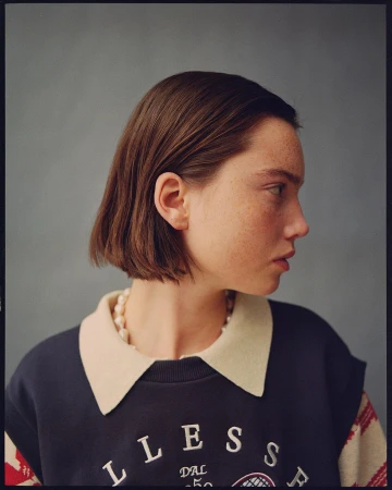 Profile of an adult woman with bobbed brown hair, wearing a vintage Ellesse sweatshirt with cream collar and pearl necklace against grey.