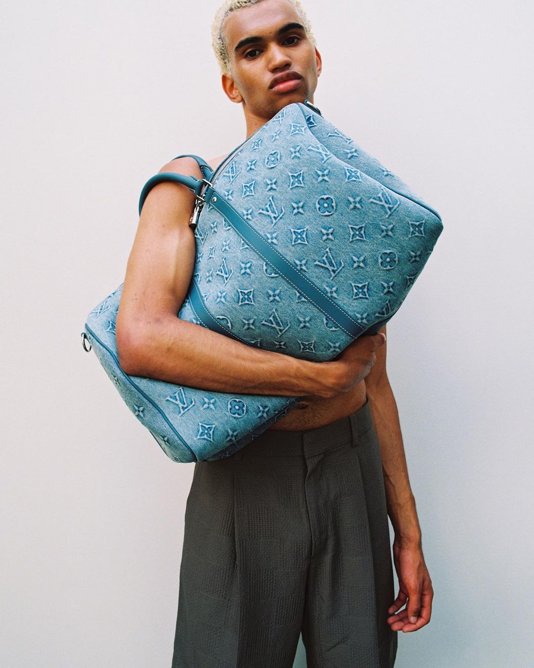 Male model poses with Louis Vuitton blue monogrammed textile travel bags against a white background in a studio setting.