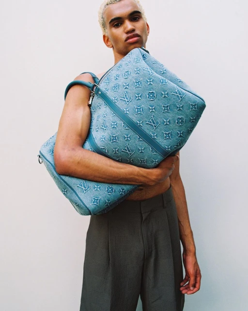 Male model poses with Louis Vuitton blue monogrammed textile travel bags against a white background in a studio setting.
