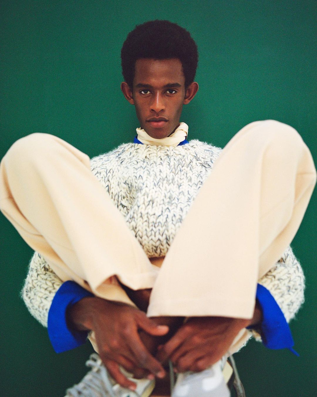 Adult male seated indoors, wearing a neutral sweater and pants, posing in front of a green background, fashion portrait.