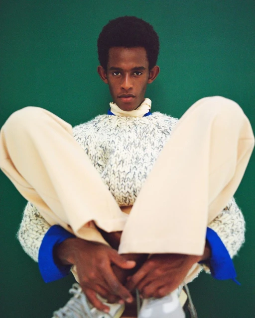 Adult male seated indoors, wearing a neutral sweater and pants, posing in front of a green background, fashion portrait.