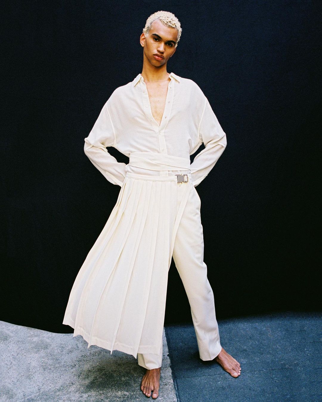 Androgynous model poses in a bright studio wearing an off-white outfit including a long-sleeved shirt, pants, and a draped skirt.