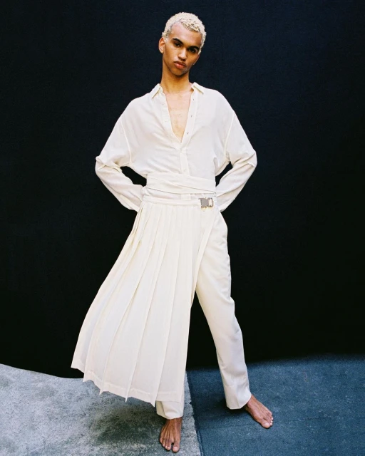 Androgynous model poses in a bright studio wearing an off-white outfit including a long-sleeved shirt, pants, and a draped skirt.