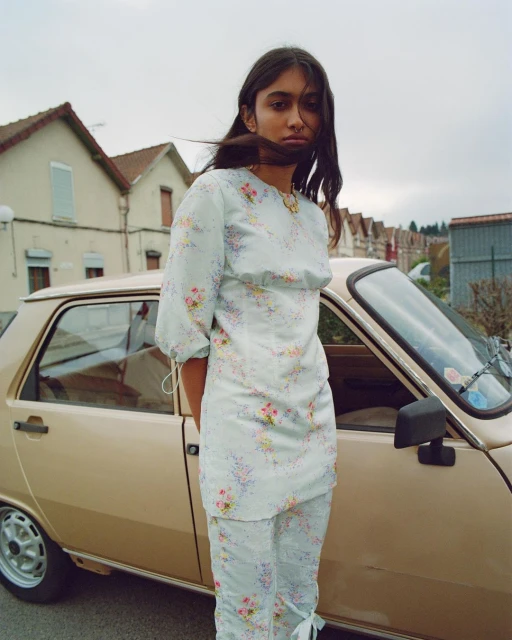 Woman in floral two-piece outfit standing next to vintage car in residential neighborhood with overcast sky and residential houses.