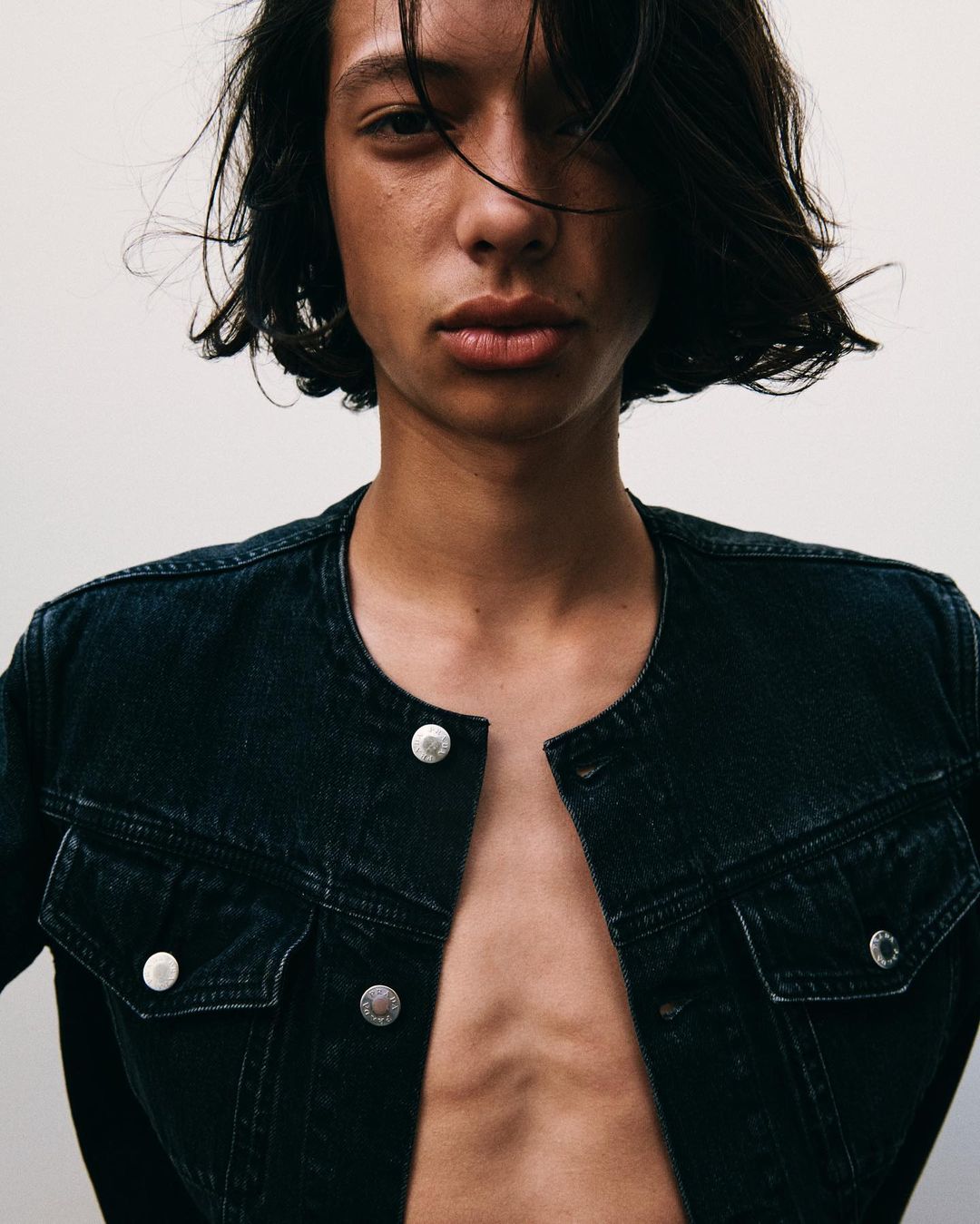 Close-up studio portrait of a slender young adult with dark hair wearing an unbuttoned dark denim jacket, showcasing minimalist fashion.