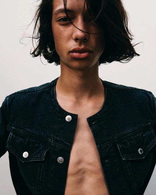 Close-up studio portrait of a slender young adult with dark hair wearing an unbuttoned dark denim jacket, showcasing minimalist fashion.