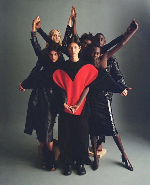 Group shot of seven diverse models in a studio setting wearing dark clothing; a model in the center wears a red, heart-shaped top.