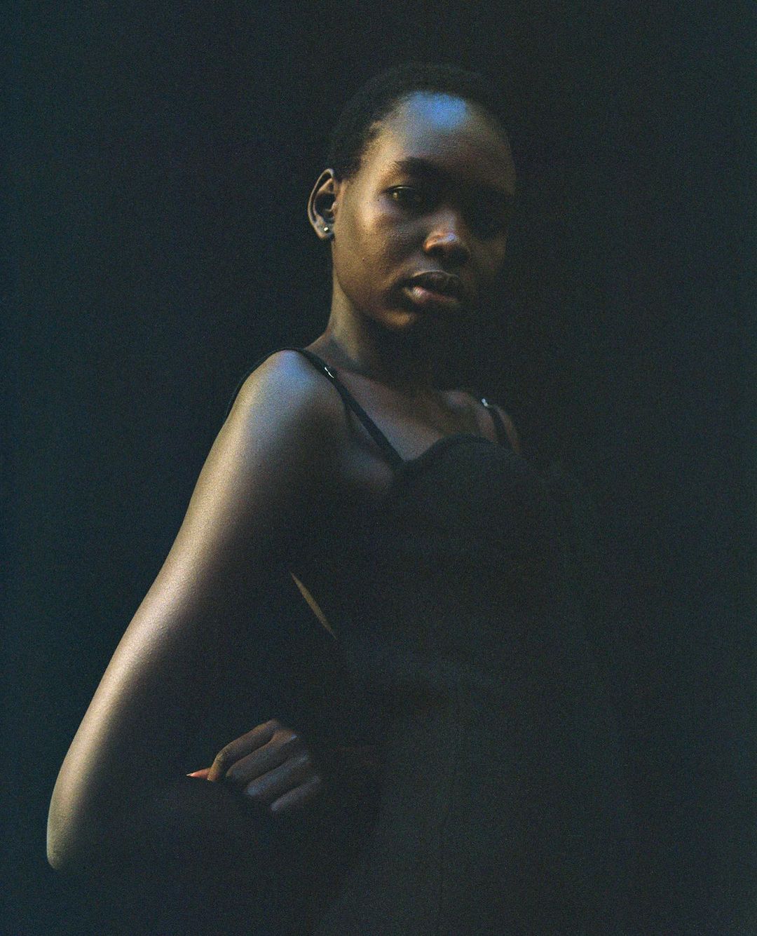 Close-up studio portrait of a young Black woman in a black top with a dark background, gazing intently at the camera with a hand on her hip.