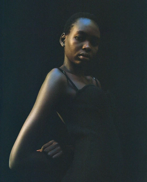 Close-up studio portrait of a young Black woman in a black top with a dark background, gazing intently at the camera with a hand on her hip.