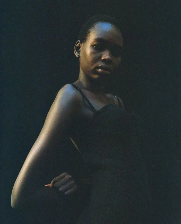 Close-up studio portrait of a young Black woman in a black top with a dark background, gazing intently at the camera with a hand on her hip.