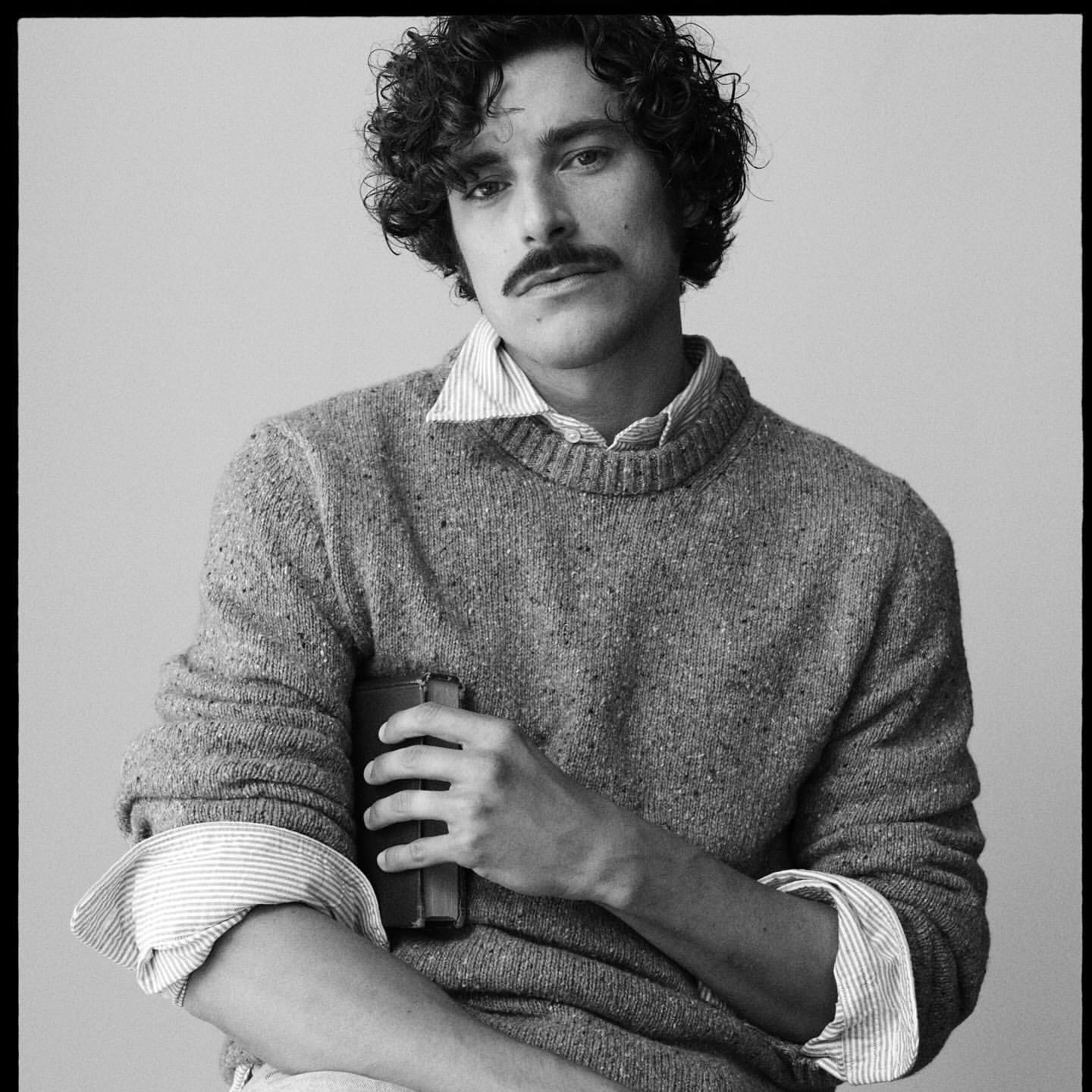 Portrait of a man with a mustache wearing a textured knit sweater holding a book, photographed by Arthur Delloye in Paris.