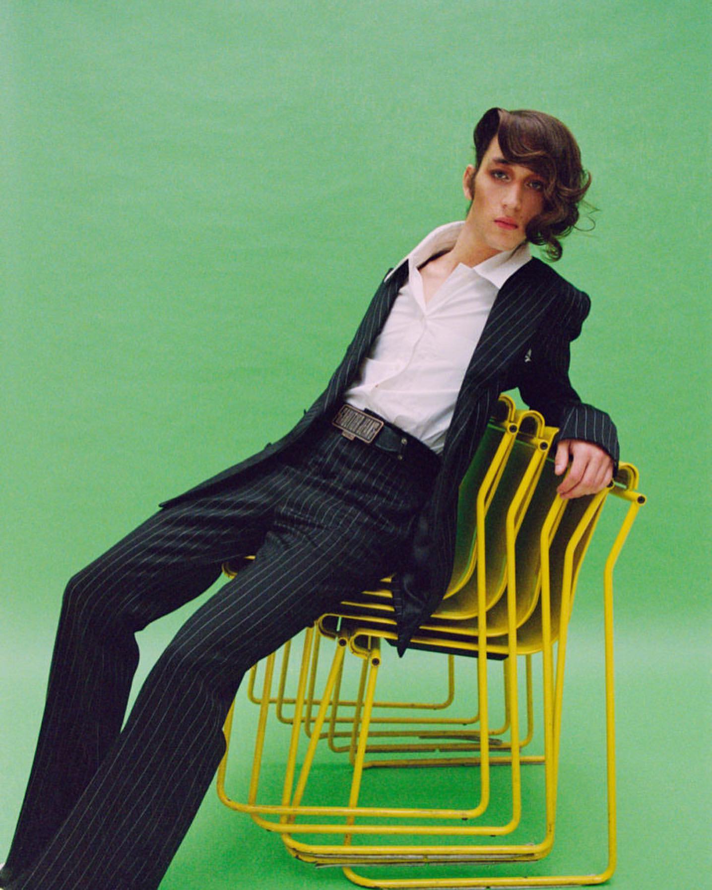 Fashion editorial shot of a slender adult male in a pinstripe suit, white collared shirt, sitting on stacked yellow chairs against green backdrop.
