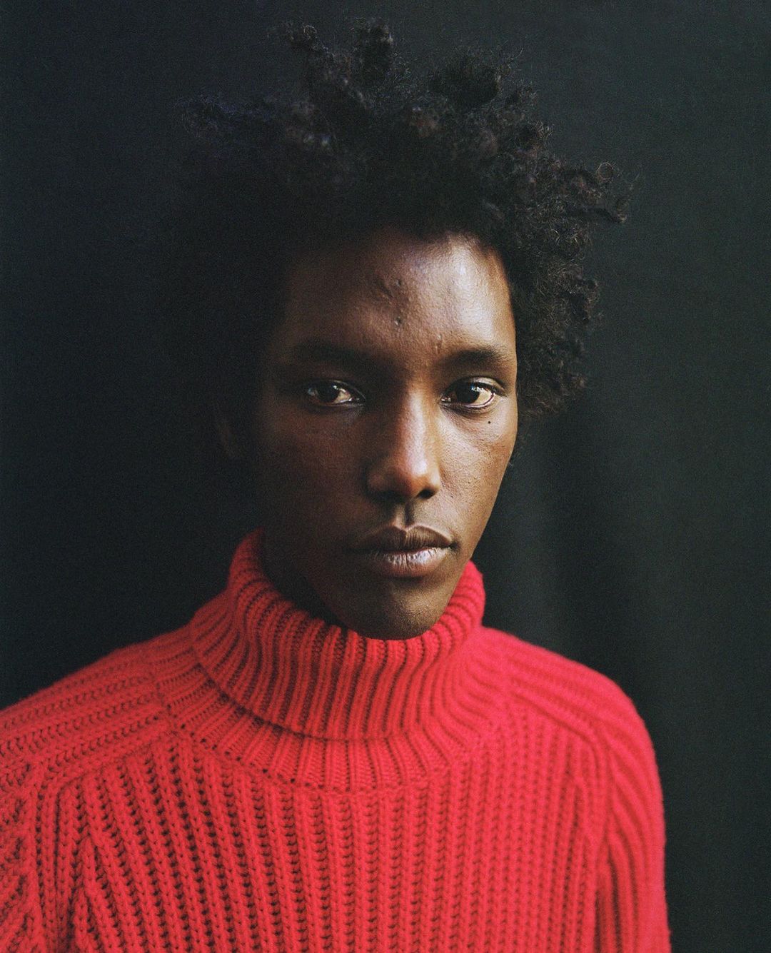 Close-up portrait of an adult male with dark skin and curly hair wearing a bright red turtleneck sweater against a dark background.