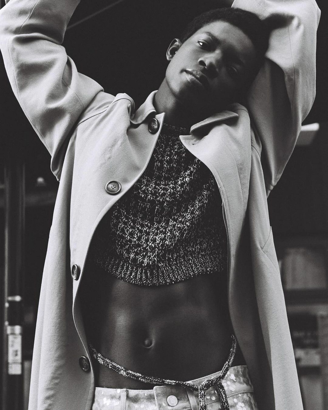 Black and white portrait of a Black male model in a cropped sweater and trench coat against an indoor background with neutral tones.