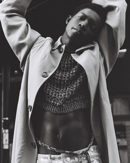 Black and white portrait of a Black male model in a cropped sweater and trench coat against an indoor background with neutral tones.