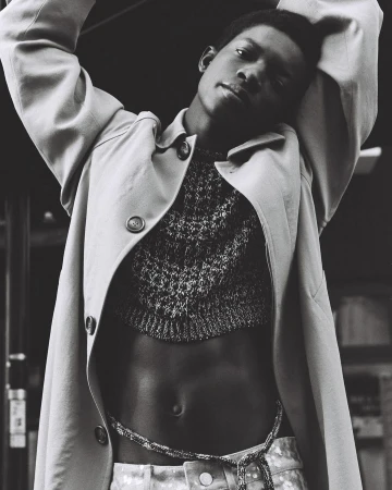 Black and white portrait of a Black male model in a cropped sweater and trench coat against an indoor background with neutral tones.