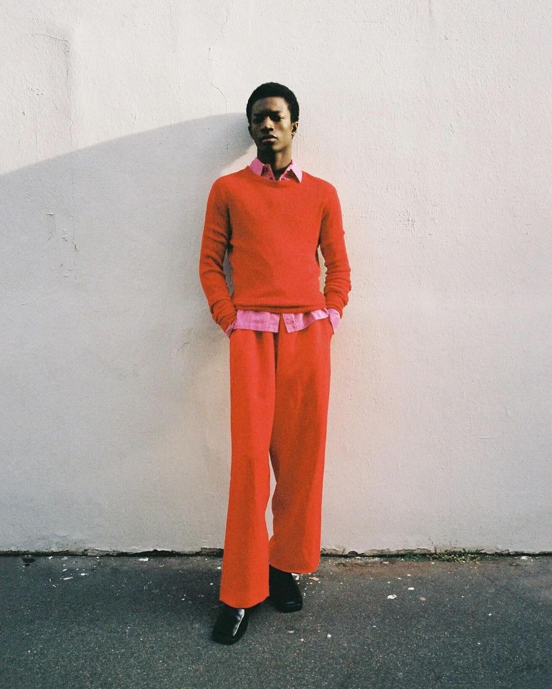 Young adult man in red sweater and pants stands against a textured wall, bathed in natural light casting shadows in an outdoor setting.