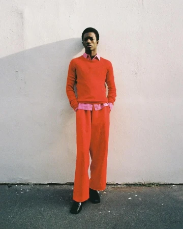 Young adult man in red sweater and pants stands against a textured wall, bathed in natural light casting shadows in an outdoor setting.