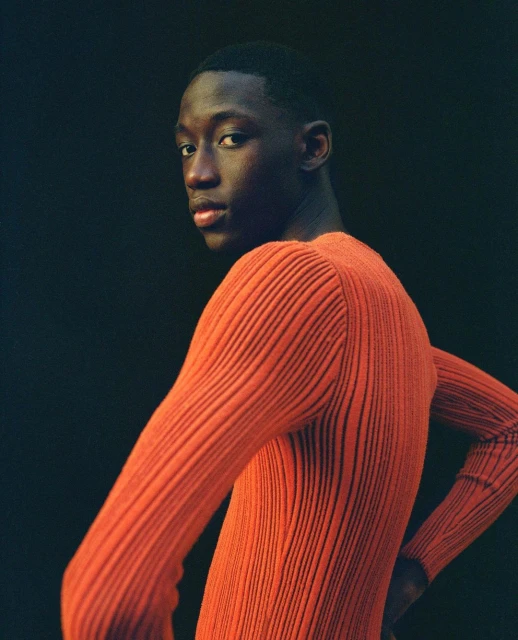 Portrait of a young adult male with dark hair and skin, wearing a ribbed orange long-sleeve shirt, posing in front of a dark background.
