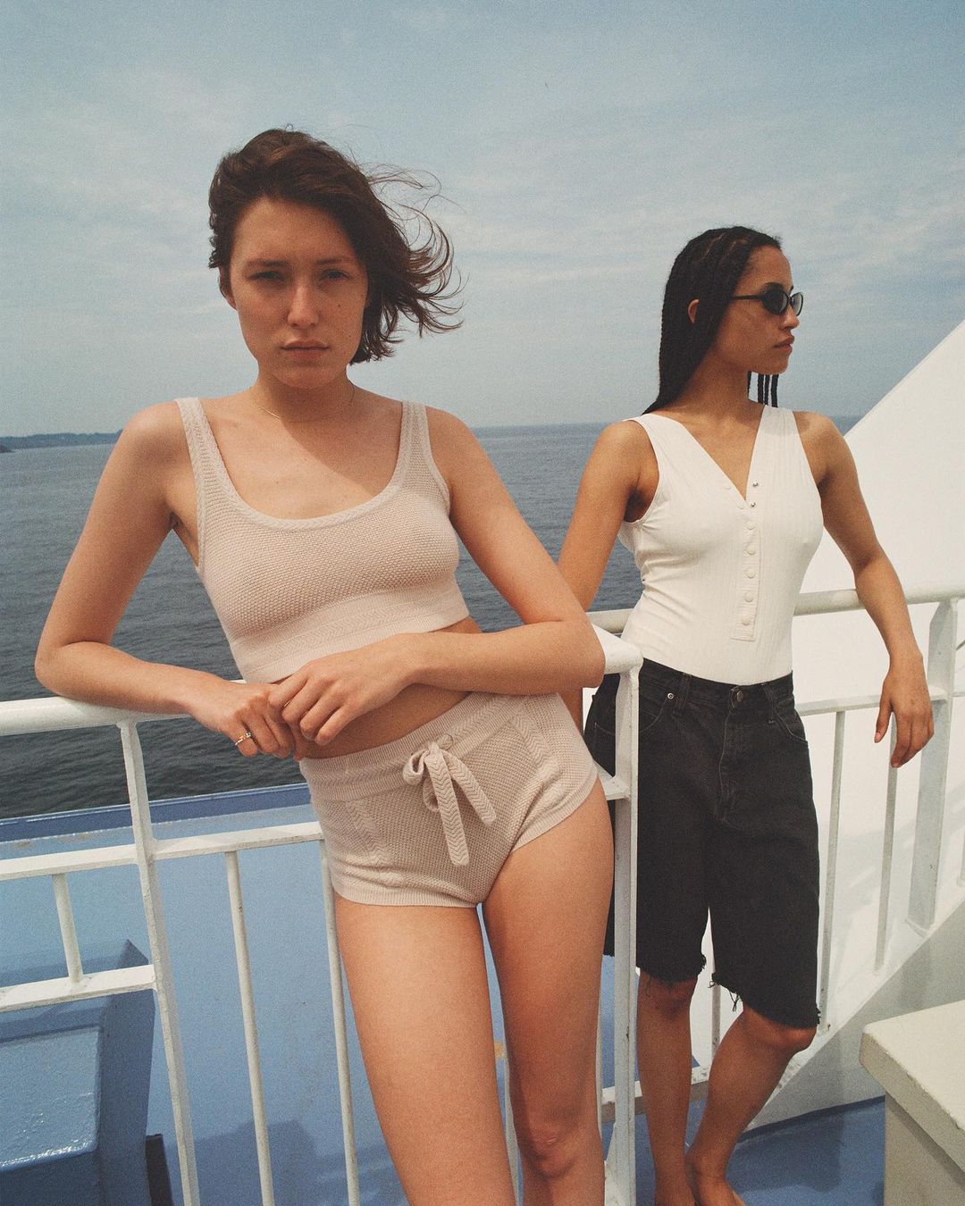 Two women model summer fashion on a boat, wearing casual tops and shorts, with the sea and sky providing a bright, nautical backdrop.