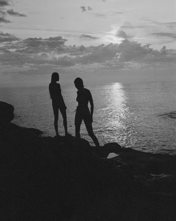 Two silhouetted figures standing on a rocky coast overlooking a calm ocean under a cloudy sunset sky.