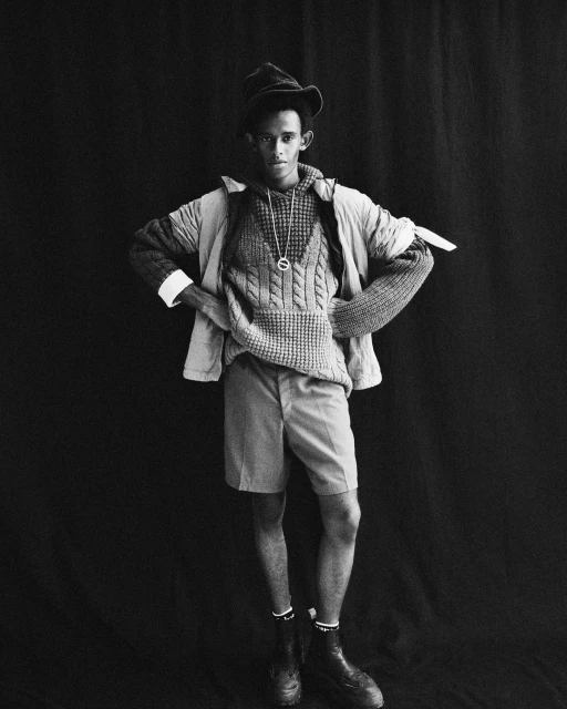 Monochrome portrait of a Black male model wearing a hat, sweater, shorts, boots against a dark backdrop in a studio setting.